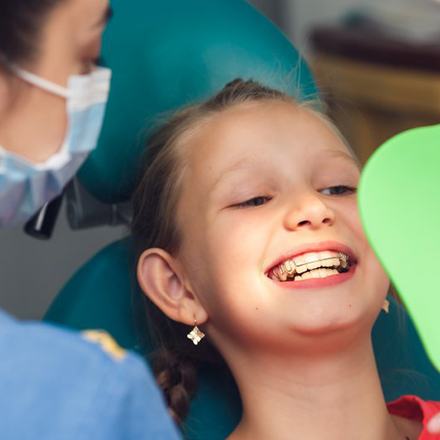 Child with braces in patient’s chair smiles at a handheld mirror